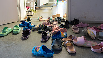 Zahlreiche bunte Kinderschuhe liegen verstreut im Eingangsbereich eines Raumes vor einem Regal mit weiteren Schuhen.