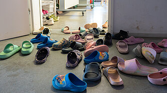 Zahlreiche bunte Kinderschuhe liegen verstreut im Eingangsbereich eines Raumes vor einem Regal mit weiteren Schuhen.