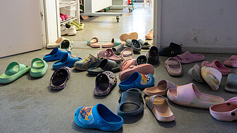 Zahlreiche bunte Kinderschuhe liegen verstreut im Eingangsbereich eines Raumes vor einem Regal mit weiteren Schuhen.