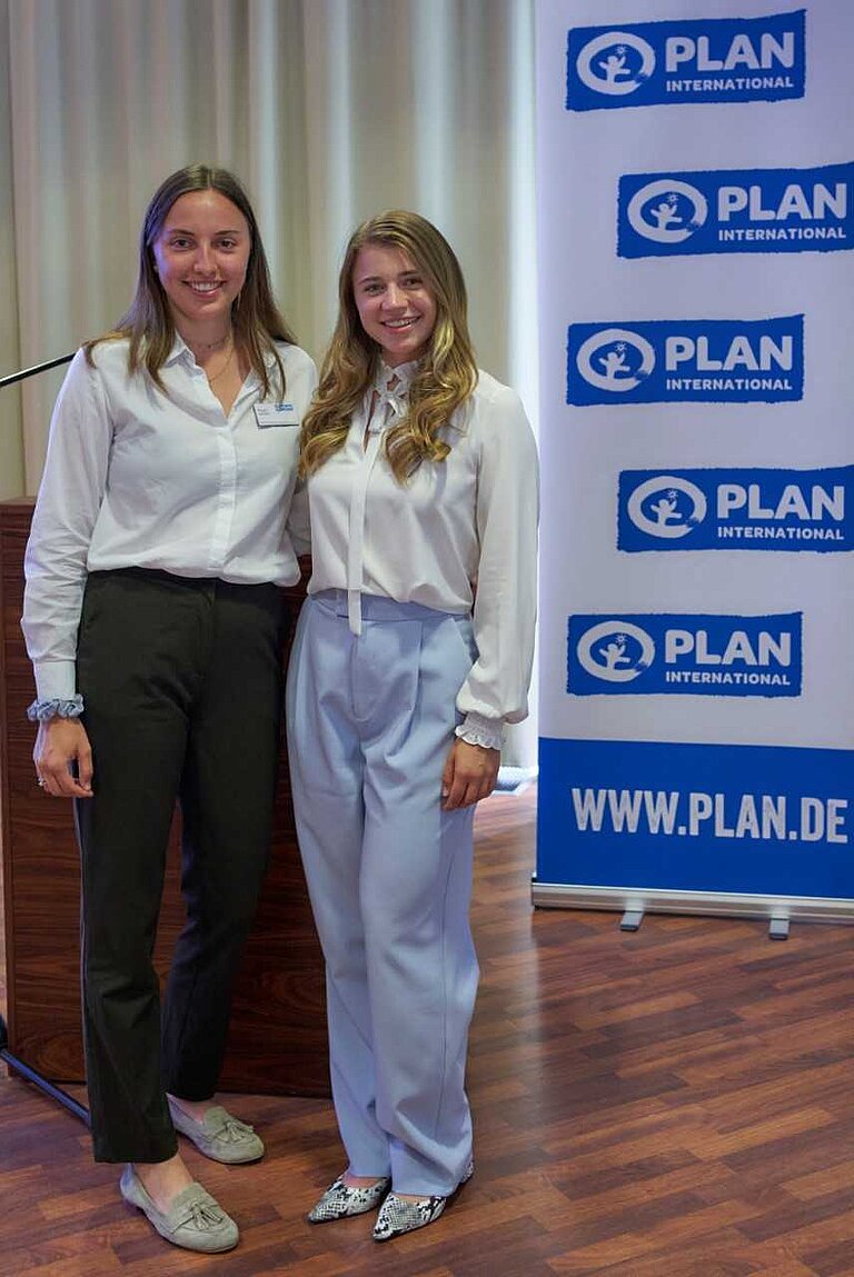 Zwei junge Frauen stehen in schicker Kleidung neben einem Plan International-Banner.