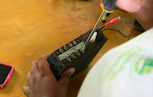 Nahaufnahme von Händen, die mit einem Schraubenzieher an einem kleinen elektronischen Bauteil arbeiten; Messkabel und ein Smartphone liegen auf einem Tisch, der als Übungsfläche dient.