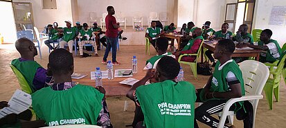 Mehrere Männer sitzen auf Plastikstühlen an Tischen in einem großen Raum. Viele tragen grüne Westen, auf denen "Papa Champion" aufgedruckt ist. In der Mitte steht eine Person, die zu der Gruppe spricht. 