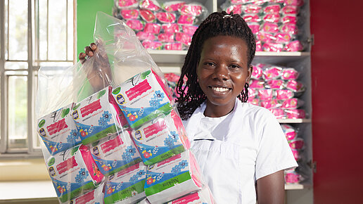 Eine junge Frau schaut in die Kamera und zeigt eine Tüte mit Menstruationsartikeln.
