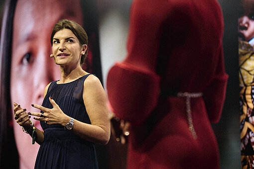 Eine Frau in einem eleganten Abendkleid hält eine Rede ; im Hintergrund ein Bild mit einem Kindergesicht.