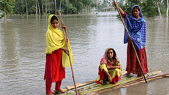 Drei Mädchen in Bangladesch überqueren das Hochwasser auf einem Floß aus Bananenstauden