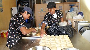 Zwei Jugendliche formen in einer Bäckerei in Ecuador Teig zu Brötchen. Auf dem Edelstahltisch liegen bereits viele geformte Teigstücke bereit zum Backen.
