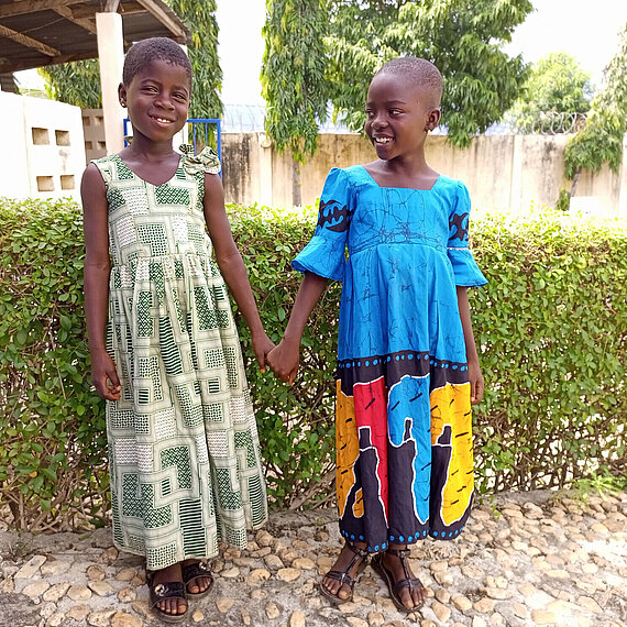  Zwei Mädchen in bunten Kleidern halten sich an den Händen und lächeln – Aufnahme in einer Schule in Togo.