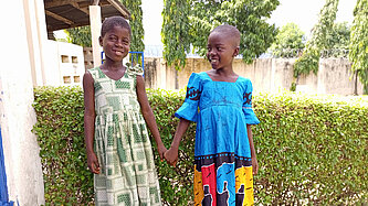  Zwei Mädchen in bunten Kleidern halten sich an den Händen und lächeln – Aufnahme in einer Schule in Togo.
