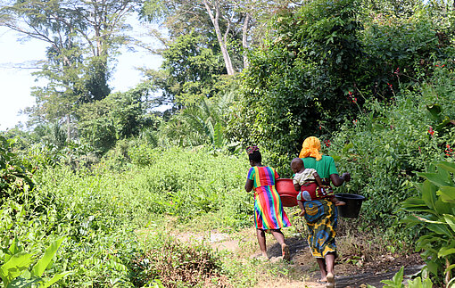 Zwei liberische Frauen laufen mit Wassereimern auf dem Kopf durch den Wald. Eine von ihnen trägt ein Kind auf dem Rücken