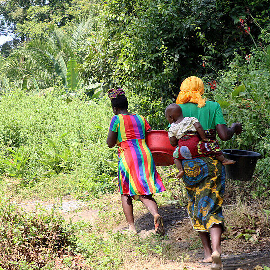 Zwei liberische Frauen laufen mit Wassereimern auf dem Kopf durch den Wald. Eine von ihnen trägt ein Kind auf dem Rücken