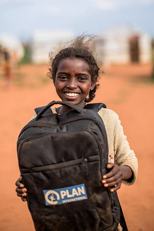 Ein äthiopisches Mädchen hält lachend einen Schulrucksack mit Plan-Logo in die Kamera