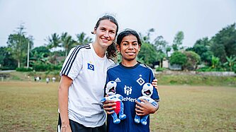 Eine junge Frau mit weißem HSV-Trikot steht zusammen mit einem dunkelhaarigen kleinen Mädchen in dunkelblauem HSV-Trikot zusammen.