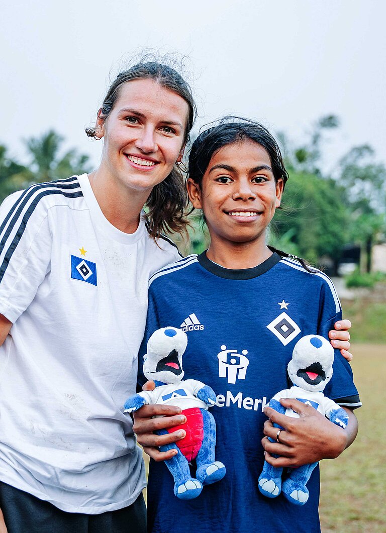 Eine junge Frau mit weißem HSV-Trikot steht zusammen mit einem dunkelhaarigen kleinen Mädchen in dunkelblauem HSV-Trikot zusammen.