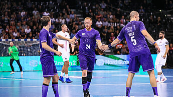 Drei Handballspieler stehen auf dem Handballfeld. Sie tragen lila Trikots mit dem Girls Get Equal-Logo.