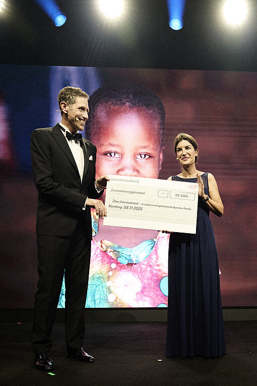 Eine Frau und ein Mann in eleganter Abendkleidung halten lächelnd einen Scheck über 92.000 Euro in die Kamera.