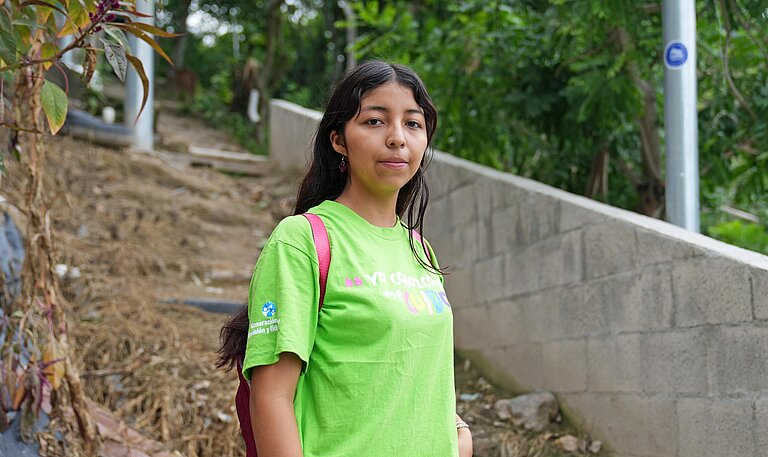 Eine junge Frau mit grünem T-Shirt steht an einem Trampelpfad im Unterholz