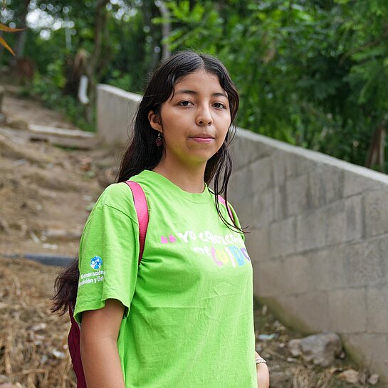 Eine junge Frau mit grünem T-Shirt steht an einem Trampelpfad im Unterholz