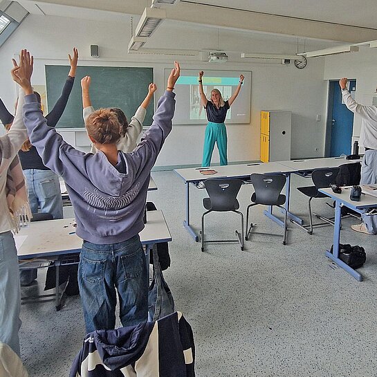 Eine Frau steht in einem Klassenzimmer vor Schulkindern und macht eine Sportübung an der Tafel vor
