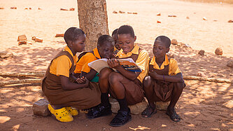 Fünf ghanaische Mädchen im Jugendalter mit Schuluniform sitzen im Schatten eines Baumes. Eines hält ein Buch in der Hand, in das alle gemeinsam reinschauen