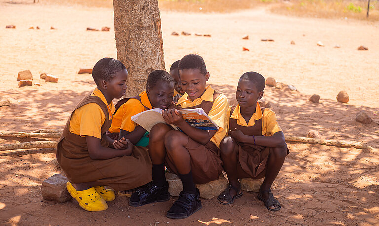 Fünf ghanaische Mädchen im Jugendalter mit Schuluniform sitzen im Schatten eines Baumes. Eines hält ein Buch in der Hand, in das alle gemeinsam reinschauen