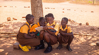 Fünf ghanaische Mädchen im Jugendalter mit Schuluniform sitzen im Schatten eines Baumes. Eines hält ein Buch in der Hand, in das alle gemeinsam reinschauen