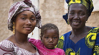 Drei Generationen afrikanischer Frauen, Mutter, Tochter und Großmutter, lächeln vor einem traditionellen Lehmhaus