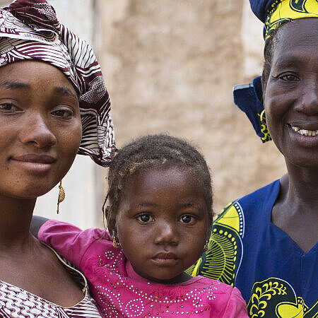 Drei Generationen afrikanischer Frauen, Mutter, Tochter und Großmutter, lächeln vor einem traditionellen Lehmhaus