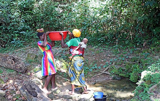 Eine liberische Mutter und ihre Tochter im Teenageralter stehen an einem Teich und heben gemeinsam einen Trog Wasser hoch