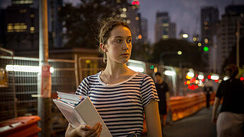 Eine junge Frau hält einen Ordner in der Hand und steht draußen in der Dunkelheit, das Umfeld sieht urban aus