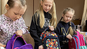 Drei Kinder, die Rucksäcke auspacken, die im Rahmen einer Nothilfeaktion im Kontext des Ukraine-Kriegs verteilt werden. Die Mädchen halten farbenfrohe Rucksäcke in den Händen.