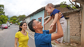 Eine Frau mit gelbem T-Shirt sieht lachend zu, wie ein Mann in blauem T-Shirt das gemeinsame Kind in den Händen hält