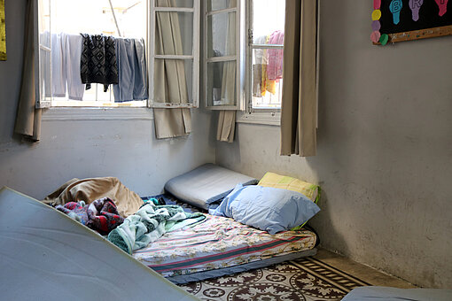 Eine einfache Unterkunft mit mehreren Matratzen und Decken, arrangiert auf dem Boden. Kleidung hängt zum Trocknen an einem offenen Fenster.
