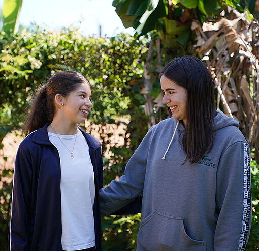 Zwei Teenager stehen in einem Garten. Eine hat ihren Arm um die andere gelegt.