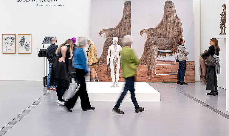 Personen laufen durch einen schneeweißen Raum, an dessen Wänden große Bilder und Fotos hängen