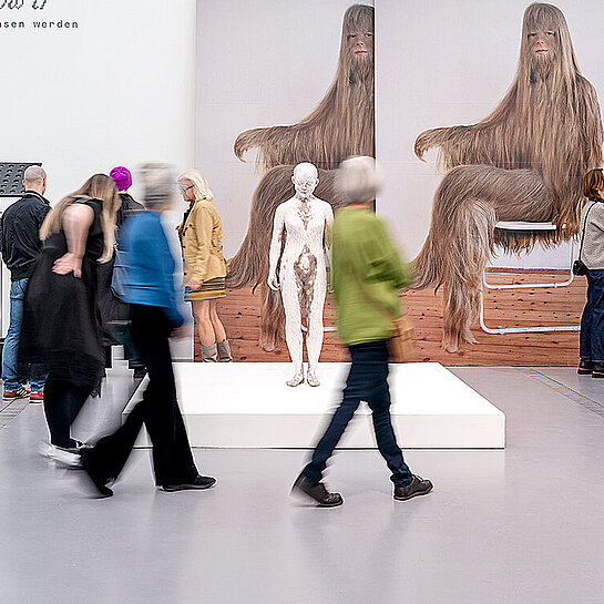 Personen laufen durch einen schneeweißen Raum, an dessen Wänden große Bilder und Fotos hängen