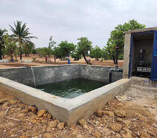 Die Bewässerungsanlage, die auch das Feld von Adjaratou inzwischen mit Wasser versorgt Ein Wasserbecken aus Stein gefüllt mit Wasser. Es ist umgeben von trockenem Boden.