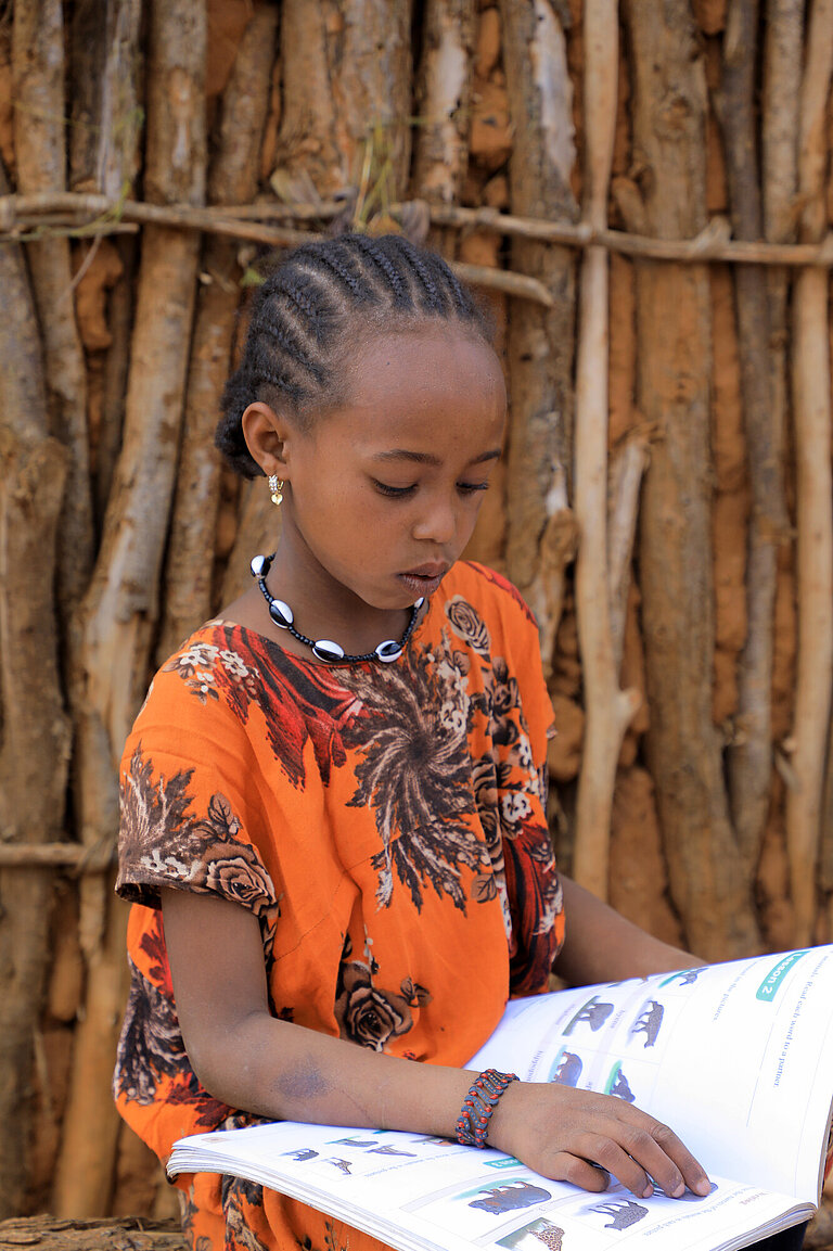 Ein Mädchen sitzt vor einer Holzhütte und liest in einem Schulbuch