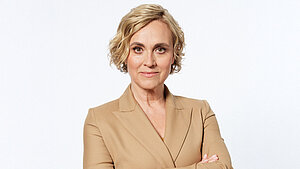 Caren Miosga steht mit verschränkten Armen vor hellem Hintergrund, trägt einen beigefarbenen Blazer und blickt selbstbewusst in die Kamera.