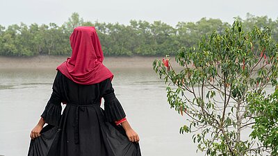 Tasnim aus Bangladesch steht in einem schwarzen Kleid mit rotem Kopftuch und schaut auf einen Fluss. Sie zeigt nur ihre Rückseite und hält mit ihren Händen ihren voluminösen Rock fest.