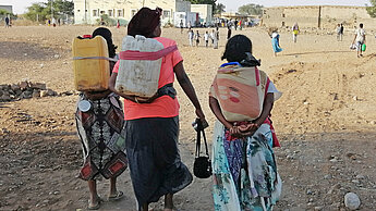 Drei Frauen, von hinten sichtbar, gehen mit schweren Wasserkanistern auf den Rücken durch eine wüstenähnliche Landschaft