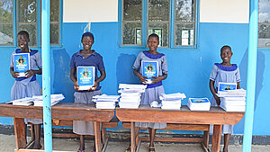 Vier ugandische Schülerinnen stehen vor einem Schulgebäude und halten Hefte mit der Aufschrift „Holiday Work“ in den Händen