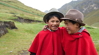 Zwei lachende Jungen in roten Ponchos und traditionellen Hüten stehen Arm in Arm in einer grünen Berglandschaft.