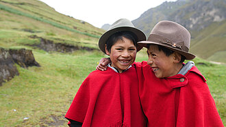 Zwei lachende Jungen in roten Ponchos und traditionellen Hüten stehen Arm in Arm in einer grünen Berglandschaft.