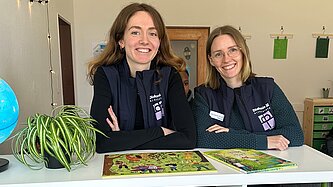 Zwei junge Frauen stehen an einem weißen Tisch und lächeln in die Kamera