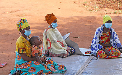 Drei Frauen sitzen auf einer Plane auf dem Boden, eine von ihnen hat ein Baby auf dem Schoß. Alle Frauen tragen einen Mund- und Nasenschutz.