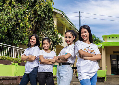 Vier Mädchen stehen nebeneinander auf einem Schulhof, alle mit verschränkten Armen und weißem T-Shirt. Sie blicken selbstbewusst und freundlich in die Kamera. Im Hintergrund sieht man ein Schulgebäude, grün gestrichene Mauern und einen großen Baum.