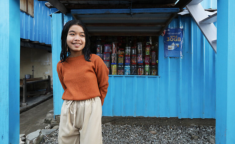 Deswira steht vor einem blauen Kiosk, sie trägt einen orangefarbenen Pullover und khaki Hosen.