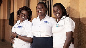 Drei Mitarbeiterinnen eines Gemeindeprojekts in Uganda stehen lächelnd vor einem Gebäude und tragen Hemden mit dem Logo von Plan International