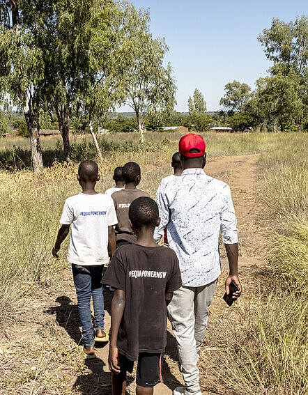 Patrick führt seine „Champions of Change“ zu der Familie eines Mädchens, das die Schule abbrechen musste Ein junger Mann in roter Mütze geht zusammen mit einer kleinen Gruppe Jugendlicher einen Feldweg entlang