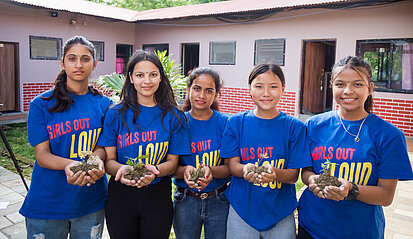 Fünf Mädchen in blauen 'Girls Out Loud'-T-Shirts halten Pflanzen in ihren Händen. Sie stehen vor einem Gebäude im Freien.